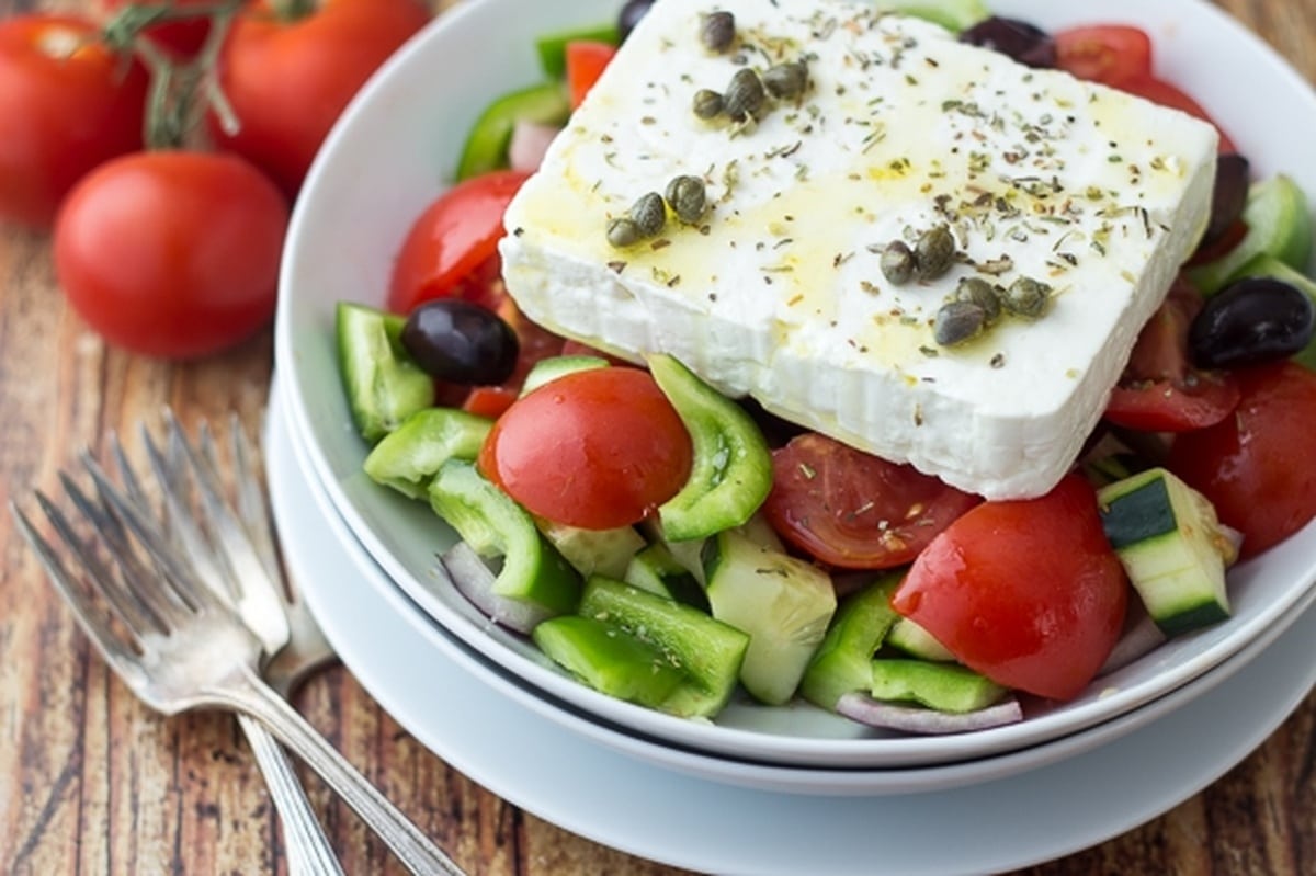 A Classic Greek Salad in a white bowl, alongside tomatoes on a table.
