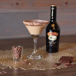 A glass of Baileys Chocolate Martini, a bottle of Baileys, and a glass with chocolate shavings on a table.