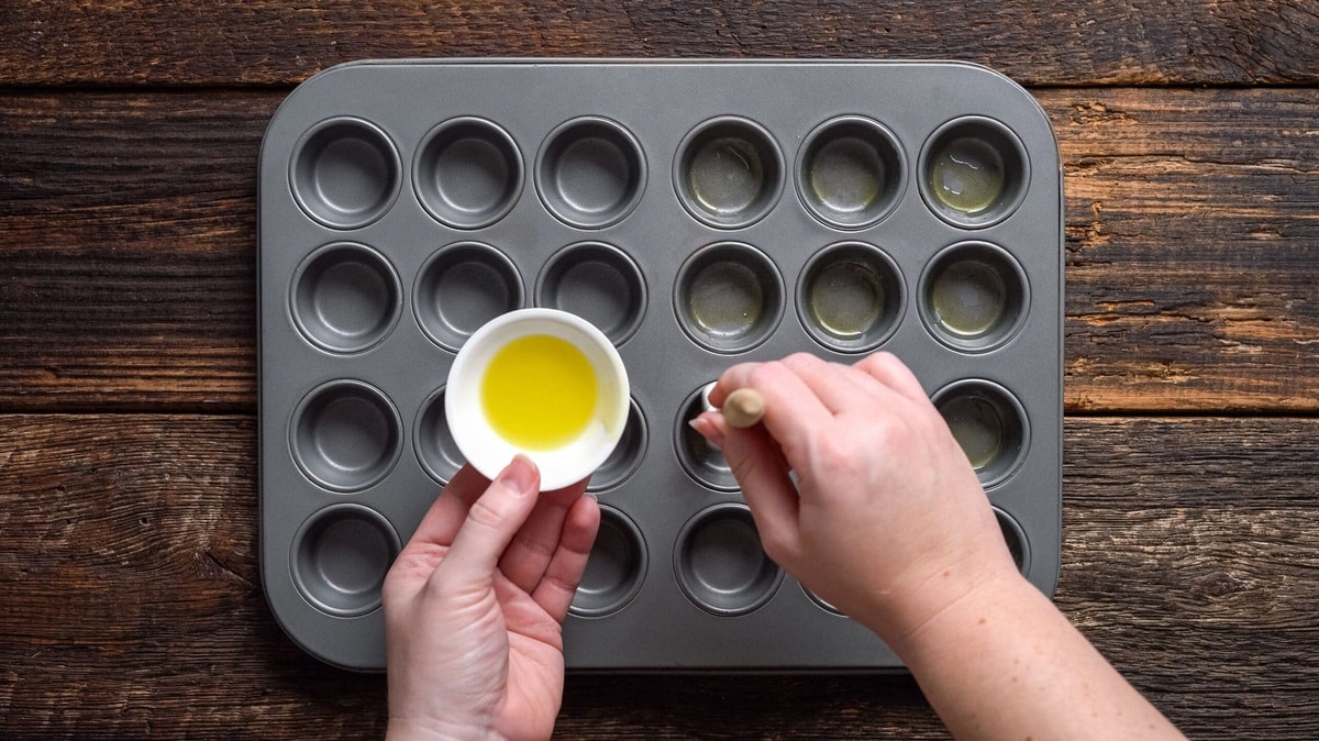 A muffin tin being greased for Brazilian cheese rolls.
