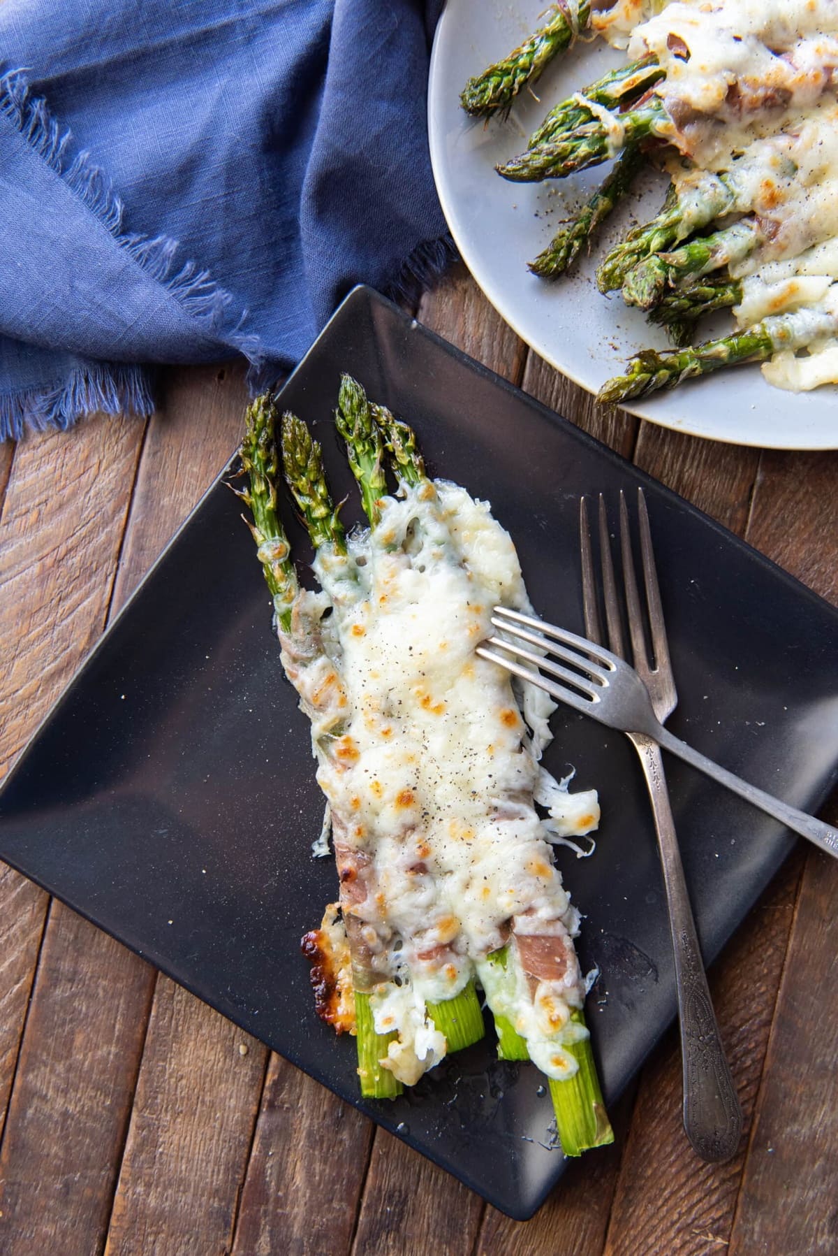 Cheesy baked asparagus on a black plate, served on a table.