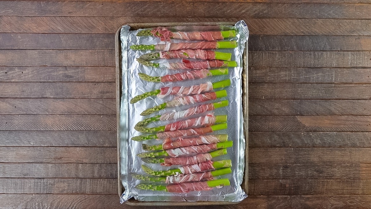 Asparagus spears wrapped with prosciutto on a baking sheet.