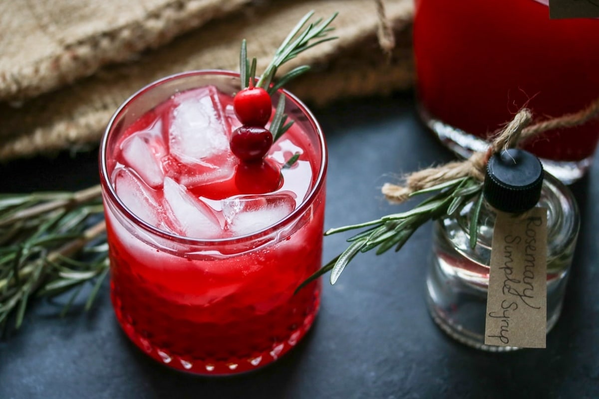 Glass of cranberry vodka spritzer with a jar of rosemary simple syrup on the side.