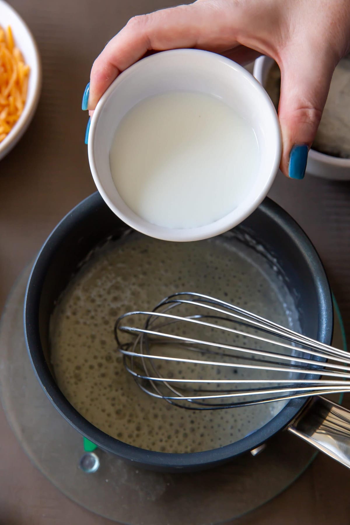 Pouring milk into the mixture while continuously whisking for a smooth and creamy texture.