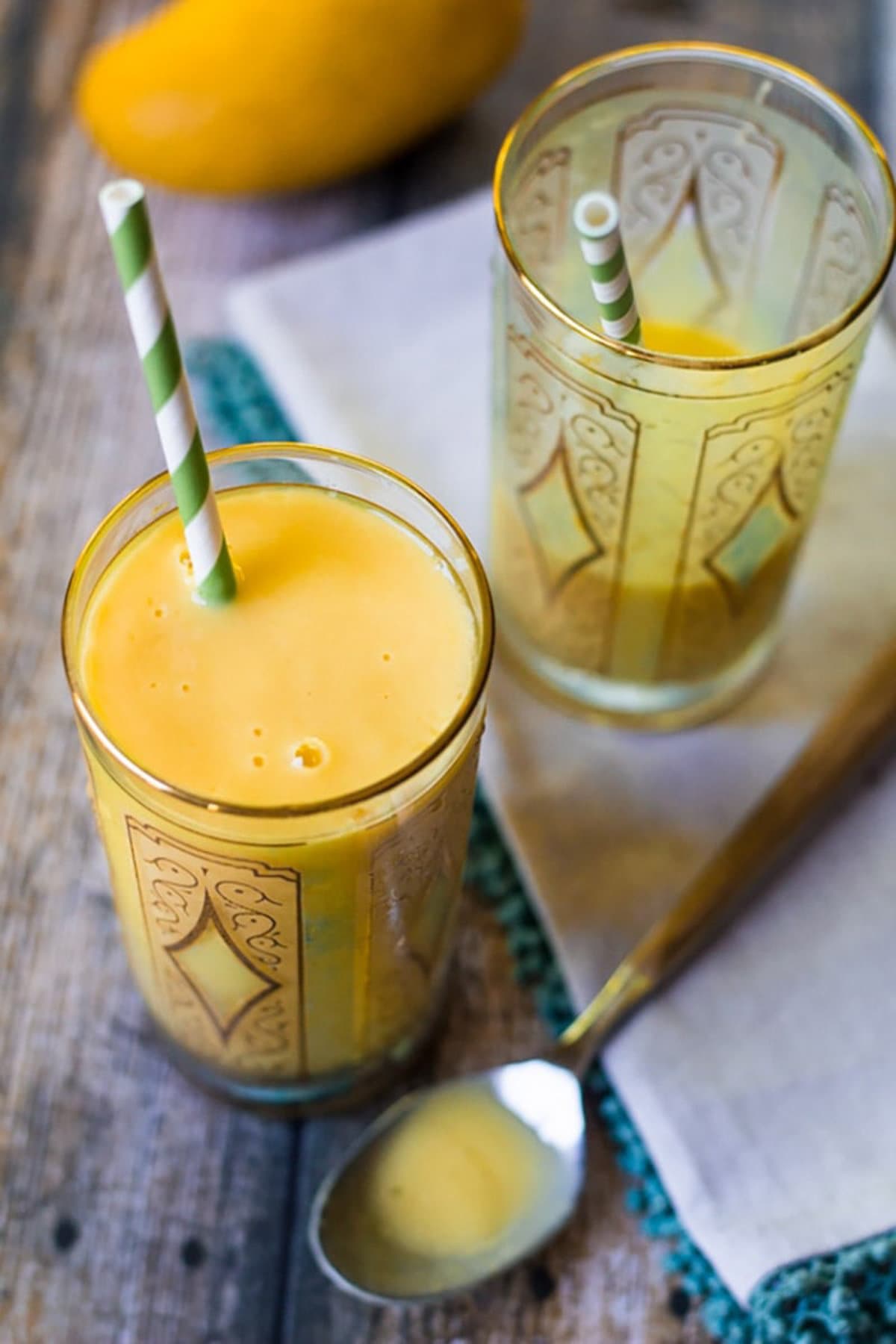 Overhead photo of two mango lasso drinks on a table with a mango and a spoon.