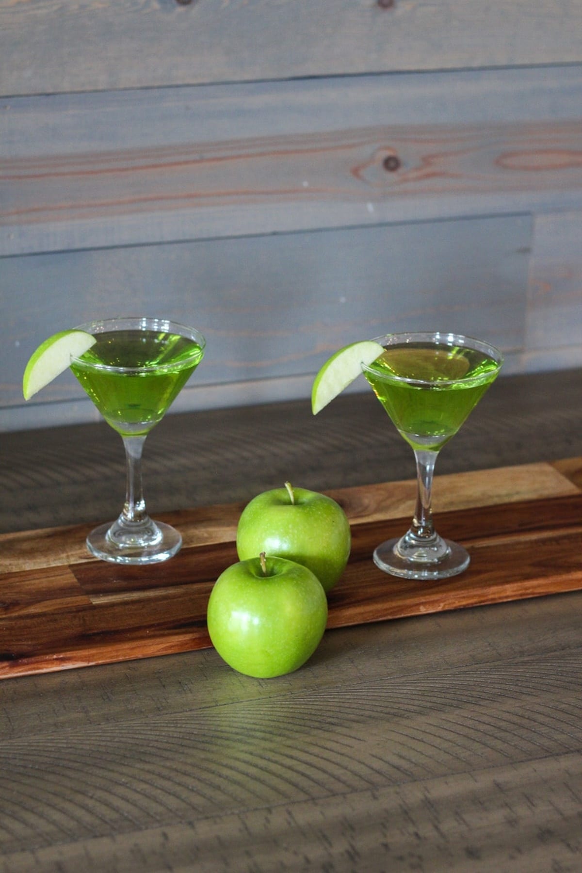 Two glasses of sour apple martini and two apples are placed on a table.